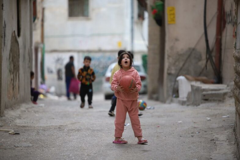 Une petite Palestinienne lève la tête, surprise, alors qu’elle joue au ballon au milieu d’une rue d’un quartier du camp de réfugiés d’Aida, près de Bethléem, en Cisjordanie occupée. (Photo : Malik Hamamra / Mondoweiss) Mai 2023.