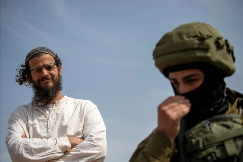 29 mars 2025. Un colon israélien qui se tient aux côtés de soldats israéliens sourit au moment où ses congénères envahissent Khirbet at-Twail, une localité située au sud de Naplouse, en Cisjordanie occupée et où vit désormais une communauté palestinienne récemment déplacée. (Photo : Avishay Mohar / ActiveStills)